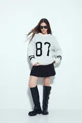 Sweater de punto color blanco con estampado numérico '87' en color negro en el frente y detalles de rayas horizontales en las mangas. Presenta cuello redondo y un corte relajado.