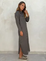 Vestido camisero largo color gris oscuro, de manga larga con presilla para sujetar el puño, cuello camisero y cierre frontal de botones. Presenta bolsillos de parche en el pecho y aberturas laterales en el ruedo.