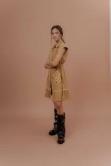 Vestido corto beige de algodón con cuello camisero y cierre frontal de botones. Presenta mangas cortas y detalles bordados a contratono en la zona del pecho y en los bolsillos, además de un volado en el ruedo.