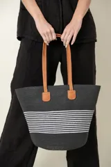 Bolso tipo tote rústico de paja trenzada color negro con rayas horizontales blancas. Tiene doble asa de cuero color marrón y división interna con cierre.