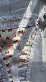 Conjunto de lencería de dos piezas confeccionado en tela acanalada blanca con estampado de pequeñas flores rosas. Incluye un soutien tipo triángulo sin aro, con detalles de encaje blanco en los bordes y una argolla metálica en el centro, y una bombacha colaless ajustable con tiras laterales y detalles de encaje.