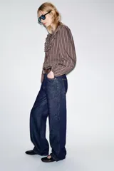 Camisa de manga larga con cuello subido, cierre frontal con botones y detalle de volantes en el pecho. Presenta un diseño a rayas verticales y puños elásticos.