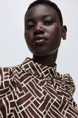 Blusa de manga larga en tejido transparente con estampado geométrico en tonos marrón y blanco. Presenta cuello elevado con frunces y puños ajustados con abertura y botón.