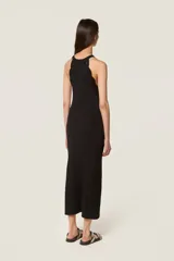 Vestido largo de punto acanalado en color negro, con diseño de sisa americana y cuello redondo decorado con una flor de crochet hecha a mano. Presenta un ribete de trenza de crochet y una abertura lateral en la parte inferior.