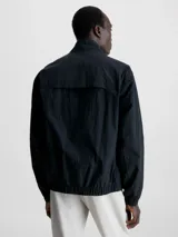 Campera negra de nailon arrugado reciclado, con cuello alto, puños y bajo elásticos, cierre con cremallera, bolsillos con cremallera en la cintura, abertura de ventilación en la espalda, forro de poliéster reciclado e insignia con el logo de goma de Calvin Klein en la manga.