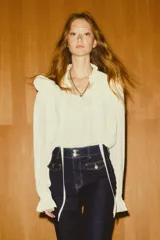 Blusa de crepé color blanco roto, con cuello alto de volado y tiras para atar en el frente. Tiene mangas largas abullonadas con puños abotonados y ribete de volado. El canesú es de doble capa con diseño en V y ribete de volado.