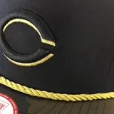 Gorra New Era 9FIFTY negra con visera de camuflaje y logo del equipo de béisbol Cincinnati Reds bordado en dorado.