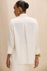 Camisa de color blanco hueso con cuello clásico, mangas tres cuartos y corte recto. Presenta un aplique decorativo frontal compuesto por piedras de colores y detalles metálicos dorados.