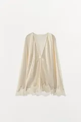 Camisa de tejido satinado con escote en pico y manga larga. Presenta un diseño holgado con cierre frontal mediante lazada y detalle de aplicación de encaje a tono en el ruedo.