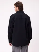 Camisa de gabardina color verde oliva, con cuello clásico, cierre frontal de botones y dos bolsillos frontales con parche. Incluye un pequeño parche con el logo de la marca en el bolsillo izquierdo.