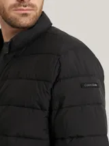 Campera acolchada negra, con cuello alto y cierre frontal de cremallera. Presenta costuras horizontales que crean un efecto acolchado y un parche con el logo de Calvin Klein en la manga izquierda.