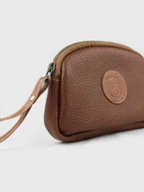 Neceser de cuero color verde oliva, con textura granulada y cierre superior. Presenta un parche circular de cuero marrón con un escudo grabado en el centro. Incluye una correa de muñeca de cuero marrón.