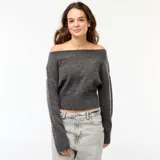 Sweater de punto gris oscuro, con escote Bardot (hombros descubiertos), manga larga y puño y ruedo acanalados. El calce es corto y holgado.