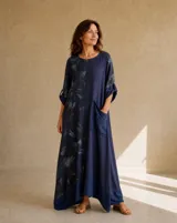 Vestido largo azul oscuro de corte holgado, con diseño asimétrico que combina un panel estampado con hojas en tonos oscuros y un panel liso con un bolsillo grande aplicado en el lateral. Tiene mangas 3/4 con presilla y cuello redondo.