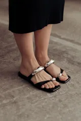 Sandalias planas negras de cuero con diseño de tiras finas. Presentan apliques metálicos plateados de forma orgánica: uno ancho sobre el empeine y otro pequeño en la tira que sujeta el dedo gordo.