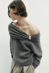 Jersey de punto color gris oscuro, con escote recto tipo off-shoulder, manga larga y detalle de aplique metálico en el hombro. Presenta acabados en rib en puños y ruedo.