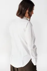 Camisa blanca de algodón con cuello clásico y cierre frontal de botones. Presenta un diseño con tablas verticales en el frente, corte redondeado en los laterales y puños anchos desmontables.