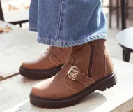 Bota de caña baja color marrón, realizada en cuero encerado, con hebillas en la caña, herrajes color níquel y cierre lateral con cremallera. El fondo está realizado artesanalmente, es de suela, y es negro. El interior está forrado en forrería vacuna.