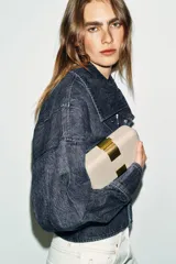 Bolso tipo clutch rígido de mano, color beige, con detalle de apliques metálicos dorados en el frente y cierre mediante clip. Incluye cadena extraíble para usar como bandolera.
