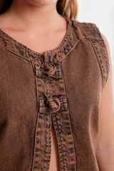 Chaleco sin mangas de denim con efecto lavado, color marrón óxido, con cuello redondo y cierre frontal con nudos decorativos.