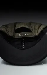 Gorra de estilo 5 paneles, color verde militar, con corona acolchada y visera negra. Presenta un pequeño parche negro con un logo blanco en forma de tijeras cruzadas.