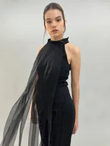 Peto negro de organza con cuello alto y cierre mediante tiras atadas al cuello, caracterizado por una capa frontal translúcida con pliegues que cae en largas tiras vaporosas.