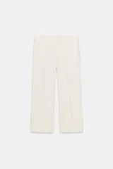 Pantalón de vestir color blanco o crema, de tiro alto y corte ancho (culotte o cropped ancho), con pliegues frontales marcados y puños anchos doblados en el bajo.