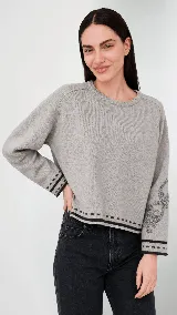 Buzo de lana gris con cuello redondo y corte holgado. Presenta un diseño bordado de arabescos en una de las mangas y detalles de rayas y puntos en contraste en los puños y el ruedo.