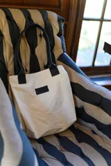 Bolso tipo tote color arena con asas de cuero sintético negro y parche con logo en el frente.