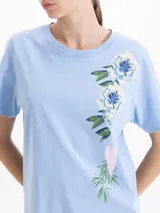 Remera de algodón con cuello redondo y mangas cortas. Presenta un diseño estampado y bordado de flores y un corazón en el frente.