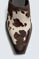 Zueco de piel con pelo, con estampado animal print tipo vaca en tonos marrones y beige. Presenta una suela de goma a contraste con un ligero tacón cuadrado y punta redondeada.