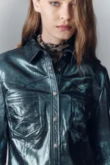 Camisa de cuero ovino verde metalizado, con cuello camisero y botones a presión con logo. Presenta un calce entallado.