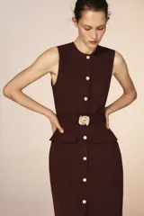 Vestido midi sin mangas de cuello redondo, con cierre frontal de botones dorados y cinturón integrado con hebilla metálica. Presenta bolsillos delanteros con solapa y una abertura en el bajo frontal.
