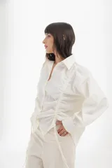 Camisa blanca de mangas largas con cuello clásico y cierre con botones. Presenta detalles de fruncido vertical en la parte delantera y cordones ajustables en los laterales. Corte holgado.