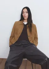 Chaqueta de ante color marrón claro, de diseño corto y recto, con cuello camisero, manga larga, dos bolsillos de parche delanteros y cierre frontal con botones.