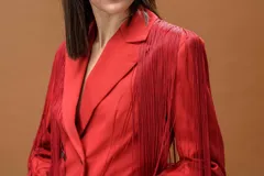 Blazer rojo de corte estructurado con solapas clásicas y cierre de doble botonadura en contraste negro. Presenta flecos largos aplicados en hombros y mangas que aportan movimiento a la prenda.