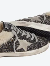 Zuecos Golden Goose modelo Super-Star Sabot, con diseño de zapatilla sin talón. Presentan exterior cubierto de glitter negro y dorado, estrella característica en el lateral, cordones y forro interior de shearling sintético.