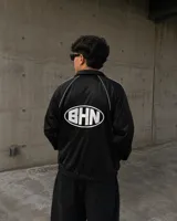 Campera deportiva de corte boxy en color negro, con diseño de cuello camisero, mangas raglán con vivos blancos y estampado circular con las siglas BHN en la espalda.