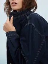 Campera de jean azul claro, estilo trucker, con cuello mao y botones metálicos plateados. Presenta costuras decorativas en el frente y mangas largas con puños doblados.