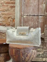 Cartera tipo tote bag de formato rectangular y alargado, confeccionada en gamuza color marrón oscuro. Presenta doble asa de hombro, solapa frontal con cierre metálico giratorio en tono dorado y costuras visibles en los bordes.