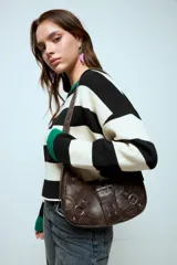 Cartera de hombro color marrón con acabado texturizado, diseño tipo hobo con hebilla decorativa frontal, remaches metálicos y argollas laterales. Cuenta con correa ajustable.