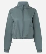 Campera cortavientos deportiva de corte cropped, color verde oscuro. Presenta capucha, cierre frontal con cremallera, mangas largas con puños elásticos y cordón ajustable en el ruedo.