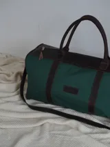Bolso tipo duffel de gran tamaño, confeccionado en lona color verde oliva con detalles y correas en cuero color suela. Cuenta con cierre superior y etiqueta de cuero con el logo de la marca.