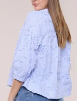 Blusa celeste a rayas con aplicaciones florales en relieve, escote en V y mangas 3/4 abullonadas con puños elásticos.