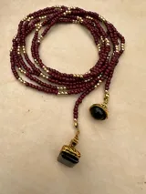 Collar largo de cuentas pequeñas en tono bordó, intercaladas con esferas doradas. Presenta dos dijes colgantes con piedras negras y detalles metálicos dorados. Su diseño permite usarlo largo o con doble vuelta.