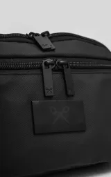 Riñonera negra confeccionada en cordura impermeable de alta resistencia. Presenta tres compartimentos principales con cierres metálicos, un bolsillo lateral adicional y una correa ajustable de alta densidad con broche de seguridad. Incluye una chapa metálica decorativa en el frente.