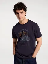 Camiseta blanca de algodón con cuello redondo y mangas cortas. Estampa rectangular con borde negro que contiene la frase "NATURAL ATMOSPHERE" en letras mayúsculas y una imagen de montañas y mar.