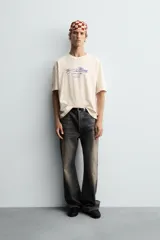 Camiseta de corte boxy fit, color beige con efecto lavado, cuello redondo y manga corta. Presenta un estampado gráfico en azul oscuro en el frente con tipografía estilo vintage.