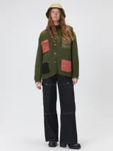 Campera tejida de punto grueso en color beige, con cuello en contraste color verde. Presenta cierre frontal con botones, bolsillos delanteros con apliques en nylon color marrón y bordado decorativo, y forro interno de nylon.