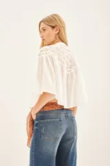 Camisa blanca de corte amplio y diseño cropped, confeccionada en algodón con detalles de encaje bordado en la parte superior y mangas acampanadas con textura rayada.